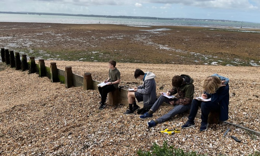 Pupils sit on a pebble beach at Hill Head collecting fieldwork data and recording observations in notebooks.